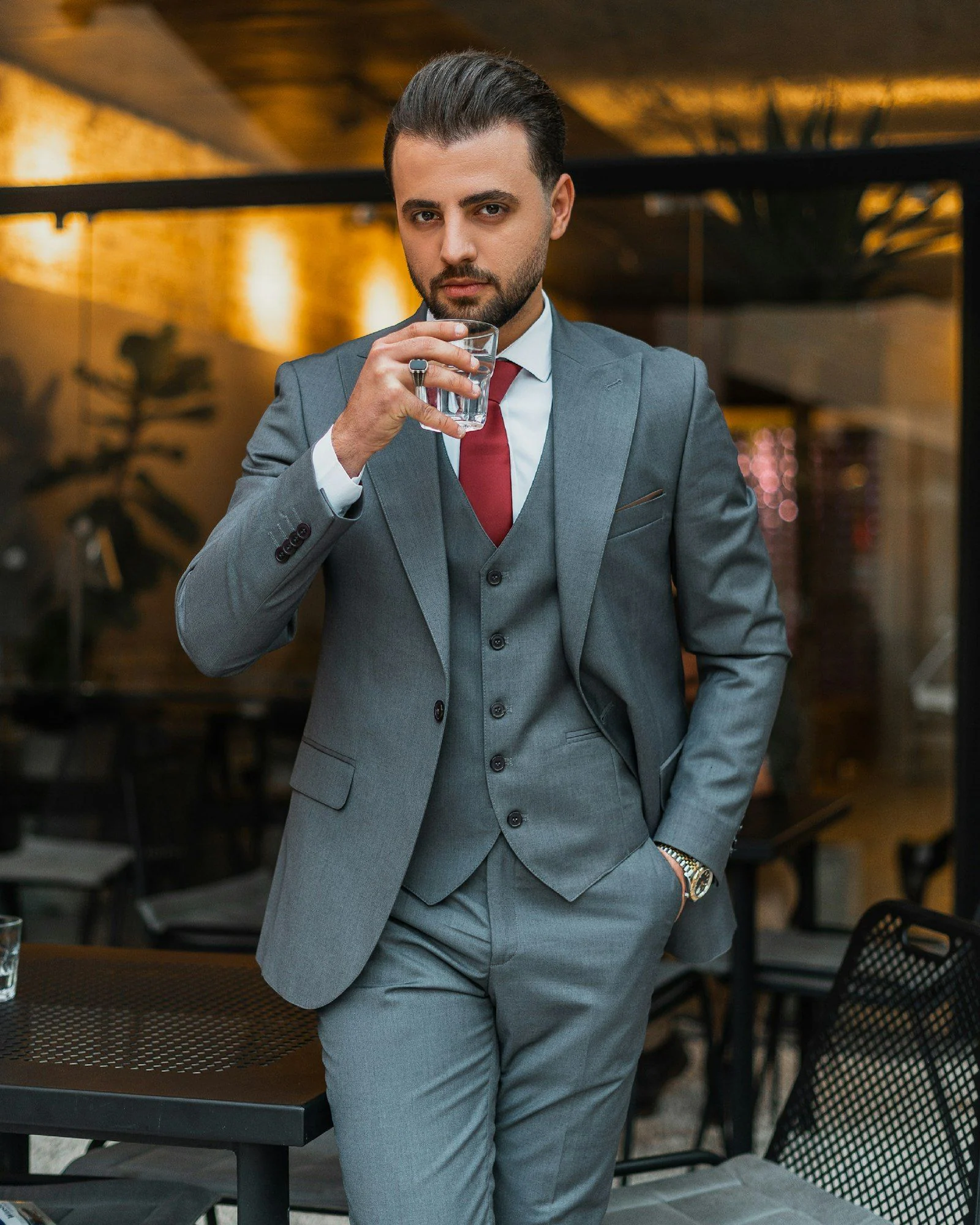 Man in a grey three-piece suit with a red tie, holding a glass